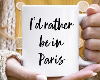 May include: White ceramic mug with the text "I'd rather be in Paris" in black script. The mug is held by a person with a cream-colored sweater in the background. The mug has a standard handle.
