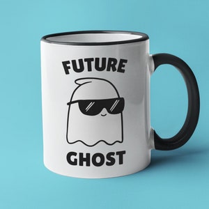May include: White ceramic mug with a black rim and handle. The mug has a black and white illustration of a ghost wearing sunglasses and the text "FUTURE GHOST".