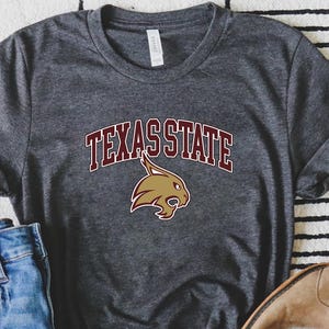 May include: A dark gray t-shirt with the text "TEXAS STATE" in maroon letters and a maroon outline. A brown bobcat mascot is centered below the text.