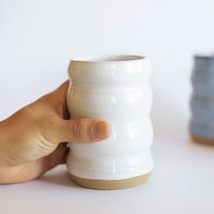May include: A white ceramic tumbler with a ribbed design is held by a hand. The tumbler has a natural clay base. Two other ceramic tumblers, one blue and one beige, are in the background. A white vase with dried flowers is also visible.