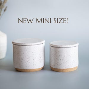 May include: Two small, white ceramic jars with lids, speckled with dark flecks. The jars have a natural, unglazed base. The text "NEW MINI SIZE!" is in brown. A white vase with dried flowers is in the background.