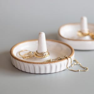 May include: Two white ceramic ring holders with a textured rim and a central post. One holder displays multiple rings and a gold necklace. The other holder has rings on the post. The holders are on a light grey surface.