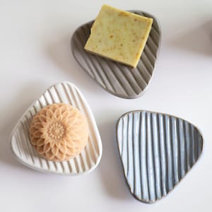 May include: Three ceramic soap dishes in a triangle shape, each with a different soap. One dish is white with a floral soap, another is gray with a square soap, and the last is blue with a ribbed design.