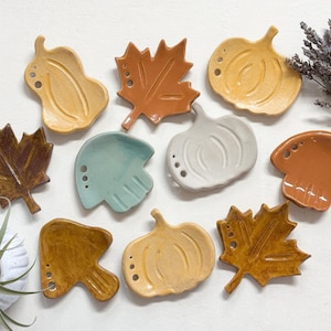 May include: A set of nine ceramic autumn-themed dishes. The dishes are shaped like leaves and pumpkins and are decorated with brown, orange, yellow, and blue glaze.