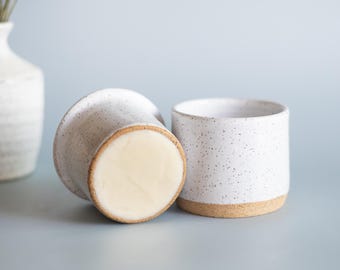 May include: Two speckled white ceramic cups with a natural tan base. One cup is upright, the other is turned over, revealing a light-colored interior. The cups are on a light blue surface, with a white vase and dried flowers in the background.