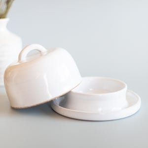Pode incluir: Manteigueira de cerâmica branca com tampa em cúpula e uma pequena pega. O prato está aberto, revelando o interior. Um vaso branco com flores secas está no fundo. O prato está sobre uma superfície azul clara.
