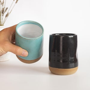 May include: Two ceramic tumblers with a cylindrical shape and a smooth, glossy finish. One tumbler is a vibrant turquoise blue with a white interior, while the other is a deep, solid black. Both feature an exposed, unglazed tan clay base.