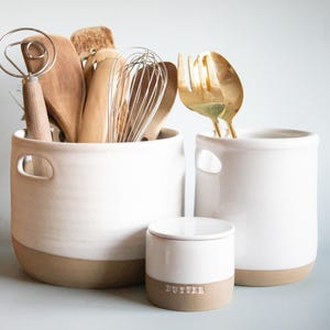 May include: White ceramic kitchen canisters with brown rims. The larger canister has two handles and holds wooden kitchen utensils. The smaller canister has a lid and says "Butter" on the front.