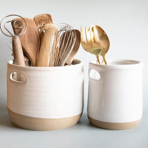 May include: Two white ceramic utensil holders with brown rims. One holder is filled with wooden kitchen utensils, including a whisk, a wooden spoon, and a dough whisk. The other holder is empty.