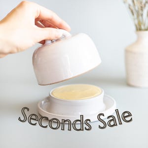 May include: A white ceramic butter dish with a dome-shaped lid and a circular base, containing a block of yellow butter. The text "Seconds Sale" is printed in a dark, stylized font across the bottom portion of the image.