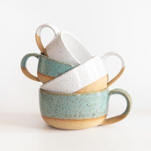 May include: A stack of three ceramic mugs with speckled glaze. The top mug is white, the middle mug is teal and white, and the bottom mug is teal. Each mug features a natural tan clay base and a curved handle, showcasing a rustic, textured finish.
