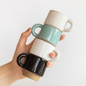 May include: A stack of four small ceramic mugs with rounded handles. The top mug is off-white, the second is a muted teal, the third is glossy white, and the bottom mug is a dark, glossy black with a visible unglazed tan base at the bottom.