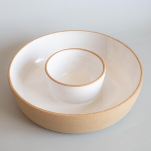 May include: A round ceramic serving dish featuring a wide outer bowl with a smaller, integrated central bowl. The interior surfaces are finished in a smooth, glossy white glaze, while the exterior walls and rims display a natural, textured tan clay.