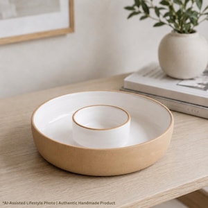 May include: A round ceramic serving dish with a tan exterior and a white interior. The dish features a smaller, integrated circular bowl in the centre, also with a white interior and a tan rim, creating a two-section design for serving food.