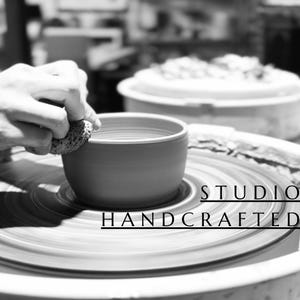 May include: A black and white image of a ceramic bowl being shaped on a pottery wheel. The text STUDIO HANDCRAFTED is visible on the right side. A textured sponge is being used to smooth the rim of the bowl while it rotates on the wheel.