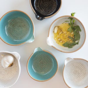 May include: Five ceramic grater bowls in various colors including teal, white, and black. Each bowl features a textured, raised center for grating. One white bowl contains garlic, oil, and herbs, while another white bowl holds a whole garlic bulb.