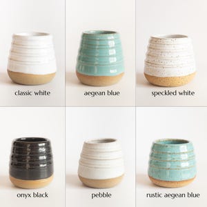 May include: A collection of six ceramic tumblers with horizontal ridges and unglazed bases. The colours include classic white, aegean blue, speckled white, onyx black, pebble, and rustic aegean blue, each labelled with its respective colour name.