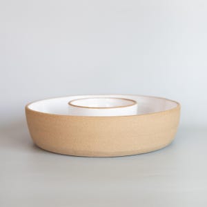 May include: A round ceramic serving dish featuring a wide outer ring and a smaller, elevated central bowl. The exterior of the dish has a textured, matte tan finish, while the interior surfaces are finished in a smooth, glossy white glaze.