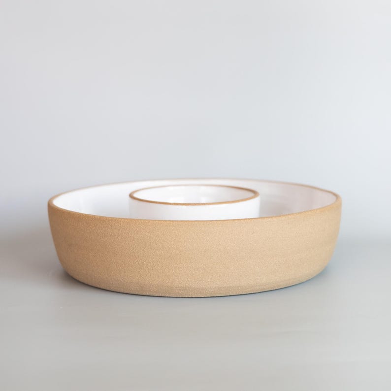 May include: A round ceramic serving dish featuring a wide outer ring and a smaller, elevated central bowl. The exterior of the dish has a textured, matte tan finish, while the interior surfaces are finished in a smooth, glossy white glaze.