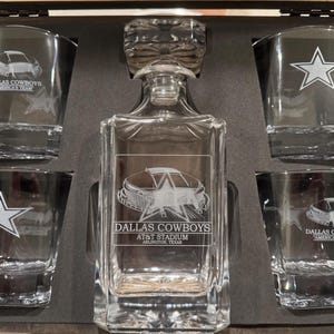 May include: A clear glass decanter and shot glass set. The decanter and shot glasses are etched with the Dallas Cowboys logo, AT&T Stadium, and a star. The set is displayed in a dark wooden box.
