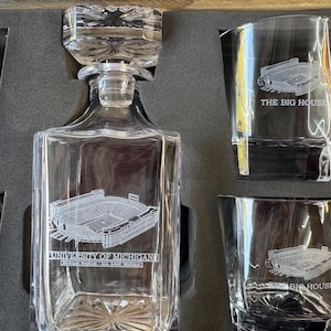 May include: A clear glass decanter set with a rectangular decanter and four matching glasses. The decanter and glasses feature an etched image of a stadium and the text "UNIVERSITY OF MICHIGAN" and "THE BIG HOUSE". The set is displayed in a black box.