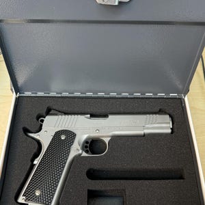 May include: A silver handgun with a black checkered grip is displayed inside a gray metal case. The case has a locking mechanism and is lined with black foam. The handgun is a 1911 R model.