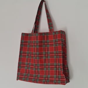 Peut inclure: Un sac fourre-tout à carreaux rouges avec des accents verts et blancs est suspendu à un mur blanc. Le sac présente un motif tartan classique, avec des poignées assorties. Le sac est en tissu robuste, idéal pour transporter des articles.