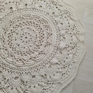 May include: Cream-colored, round crochet rug with an intricate floral and geometric pattern. The rug features a scalloped edge and openwork details, creating a textured surface. The rug is made of a soft, natural fiber.