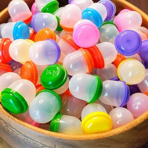 May include: A wooden bowl filled with a variety of colorful plastic capsule toys. The capsules have clear, translucent bodies and brightly colored caps in shades of pink, purple, green, orange, yellow, and blue. The bowl is made of light-colored wood.