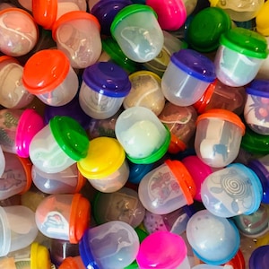 May include: A collection of colorful plastic capsule toys with various colored lids, including pink, orange, green, yellow, and blue. The clear capsules contain small toys or trinkets. The image is a close-up shot of the capsules.