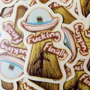 May include: A collection of stickers featuring a cartoon UFO design. The UFO is brown and beige with a blue and white eye and the words "Fucking Finally" in pink lettering. A small silhouette of a person is below the UFO.