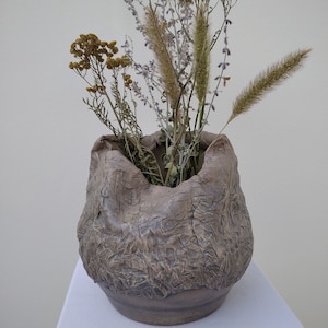 Op de afbeelding: Een bruine keramische vaas met een getextureerd oppervlak, met een boeket gedroogde bloemen met gele en paarse bloemen.