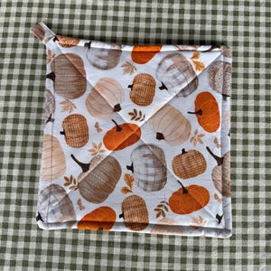 May include: A square quilted potholder featuring a white background printed with a repeating pattern of pumpkins in shades of orange, brown, and gray, accented with small brown leaves and a small fabric loop at the top corner.