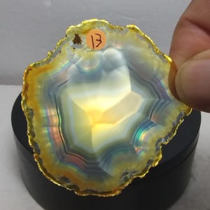May include: A translucent agate slice with concentric bands of white, yellow, and iridescent blue. The edges are finished with a metallic gold trim. A small circular orange sticker with the handwritten number 13 is attached to the top edge.