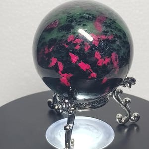 May include: A polished spherical stone featuring a deep green and black base with vibrant magenta-pink inclusions. The sphere is displayed on a silver-toned metal stand with three curved legs and intricate scrollwork details at the base.
