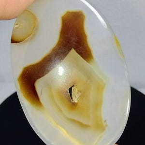 May include: An oval-shaped polished gemstone with a translucent white base featuring prominent brown and amber-colored inclusions. The stone displays natural banding patterns and a small central cavity, showcasing a smooth, glossy surface texture.