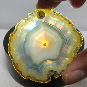 May include: A translucent, circular agate slice featuring concentric bands of white, pale blue, and yellow. The edges are trimmed in a metallic gold finish. A small circular sticker with the handwritten number 6 is attached to the top edge.
