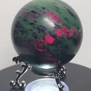 May include: A polished spherical stone featuring a deep green base with prominent inclusions of vibrant magenta and dark black mineral patterns, resting securely on a decorative silver-toned metal tripod stand with curved legs.