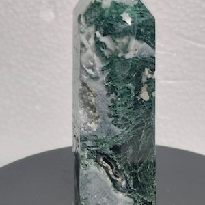May include: A polished crystal tower featuring a mix of deep forest green and translucent white mineral inclusions. The stone has a hexagonal shape with a pointed top, displaying natural internal fractures, crystalline textures, and dark green moss-like patterns throughout the structure.