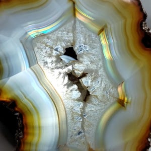 May include: A polished agate slice featuring concentric bands of amber, gold, and white colors. The center contains a crystalline geode cavity with sparkling white mineral formations. A small yellow circular sticker with the number 4 is attached.