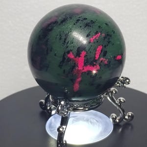 May include: A polished spherical stone featuring a deep forest green base with prominent inclusions of vibrant magenta and black mineral deposits. The sphere rests securely on a silver-toned metal stand with ornate, curved legs and a circular base.
