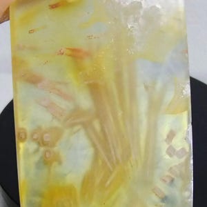 May include: A rectangular polished gemstone slab featuring a translucent pale yellow base with internal inclusions of orange and brown needle-like formations, small circular patterns, and a textured white crystalline edge at the top.