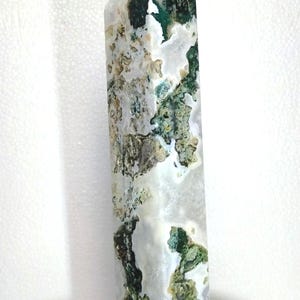 Könnte beinhalten: Ein hoher, rechteckiger Kristallpunkt mit facettierter Spitze. Der Stein weist eine Mischung aus weißen, grünen und beigen Farben mit einem moosartigen Muster auf. Der Kristall steht auf einem schwarzen Sockel.