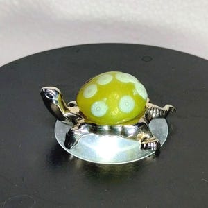 Peut inclure: Une petite figurine de tortue dotée d'un corps en métal argenté poli et d'une carapace arrondie et lisse en verre vert citron translucide. La carapace est ornée d'un motif de points circulaires blancs sur toute sa surface.