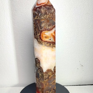 May include: A polished stone tower with a hexagonal prism shape, featuring a complex pattern of translucent white, deep orange, and dark brown mineral inclusions. The crystal point has a smooth, reflective surface and a sharp, faceted apex.
