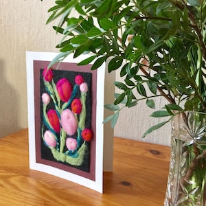 Peut inclure: Une carte de vœux faite à la main avec un motif floral en feutre. La carte a une bordure blanche et un cadre intérieur marron. Les fleurs en feutre sont dans des tons de rose, de rouge et d'orange, avec des tiges vertes et un fond noir.