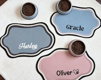Può includere: Tre tappetini sagomati per animali domestici in blu, azzurro e rosa con bordi neri. I tappetini riportano i nomi Harley, Gracie e Oliver stampati in nero o bianco. Tre ciotole in acciaio inossidabile piene di crocchette sono posizionate sopra.