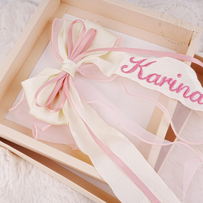 Puede incluir: Un lazo blanco y rosa con una cinta rosa que dice "Karina" bordada.