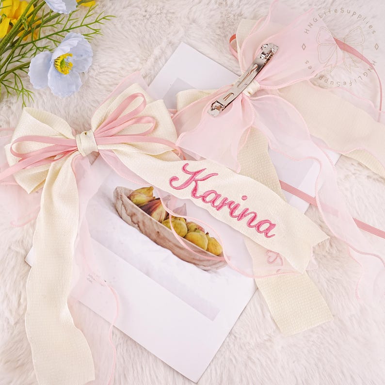 Puede incluir: Dos lazos para el pelo de cinta rosa y blanca con un nombre personalizado, "Karina", bordado en rosa en uno de los lazos.