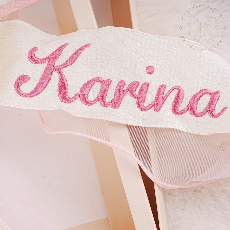 Puede incluir: Una cinta de sat&eacute;n blanca con el nombre "Karina" bordado en letras cursivas rosas.